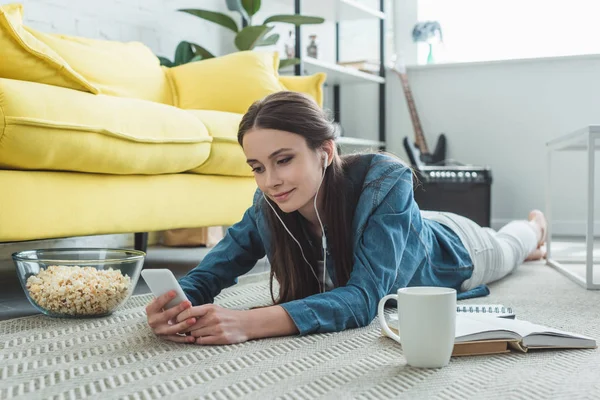 Chica sonriente en los auriculares usando el ordenador portátil mientras está acostado en la alfombra y estudiar en casa - foto de stock