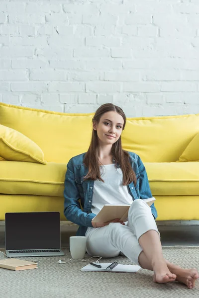 Beautiful teenage girl reading book and smiling at camera while sitting on carpet and studying at home — Stock Photo