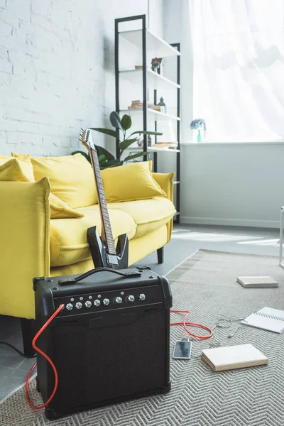 Guitarra eléctrica y altavoz con smartphone de pie en el suelo con libros cerca del sofá - foto de stock