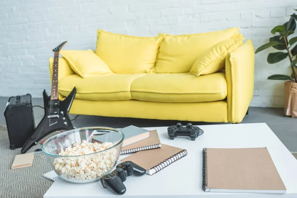 Popcorn, joysticks and copybooks on table, electric guitar near couch behind — Stock Photo