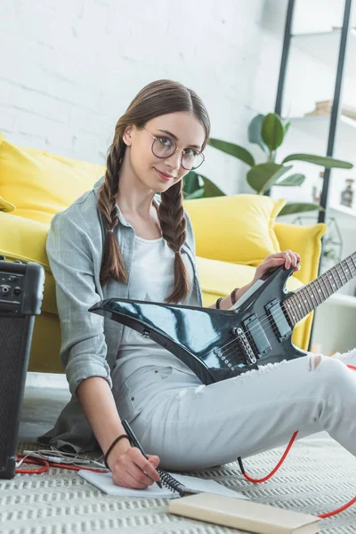 Привлекательная девушка-подросток с электрогитарой, которая пишет песни в ноутбуке, сидя на полу — стоковое фото