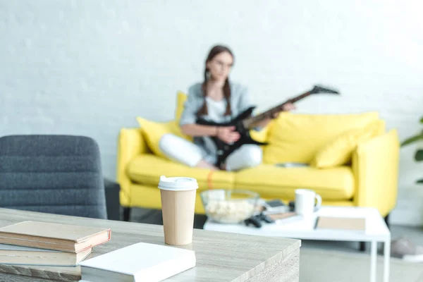 Girl playing electric guitar on sofa, boks and disposable cup of coffee on table on foreground — Stock Photo