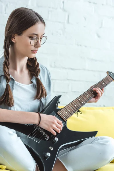 Attractive teen girl playing black electric guitar on sofa — Stock Photo