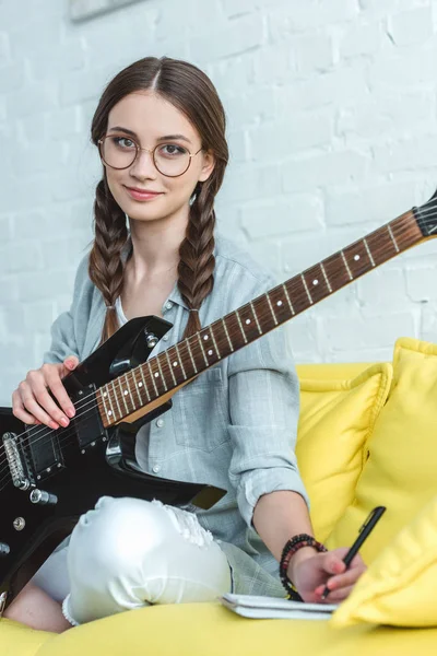 Schönes Teenie-Mädchen mit E-Gitarre schreibt Song in Lehrbuch — Stockfoto