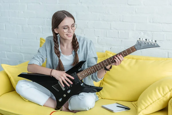 Lächelndes Teenie-Mädchen spielt E-Gitarre, während es auf gelbem Sofa mit Lehrbuch sitzt — Stockfoto