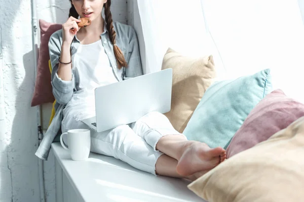 Cropped view of girl using laptop and eating cookie with coffee on windowsill — Stock Photo