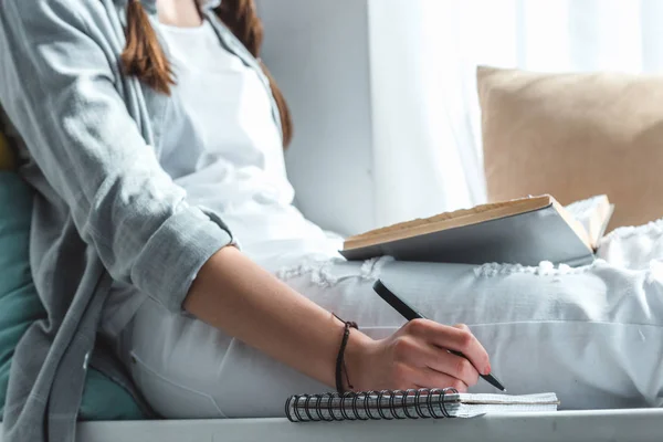 Cropped view of girl reading book and writing in copybook — Stock Photo