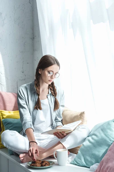 Attractive girl with braids reading book and eating cookies with coffee — Stock Photo