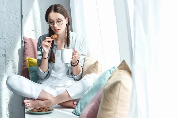 Красива дівчина їсть печиво з кавою на підвіконні з подушками — стокове фото