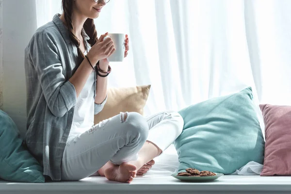 Vue recadrée de fille avec des biscuits et du café assis sur le rebord de la fenêtre avec des oreillers — Photo de stock