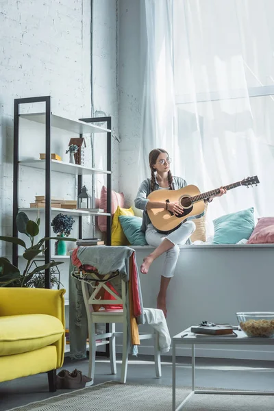 Девочка-подросток, играющая на акустической гитаре, сидя на подоконнике в гостиной — стоковое фото