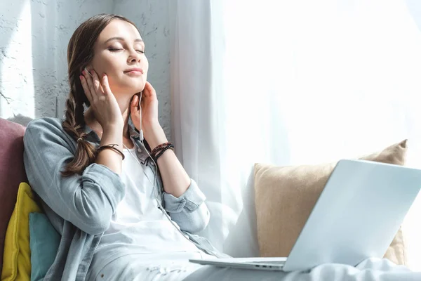 Hermosa chica adolescente usando el ordenador portátil y escuchar música con auriculares en el alféizar de la ventana - foto de stock