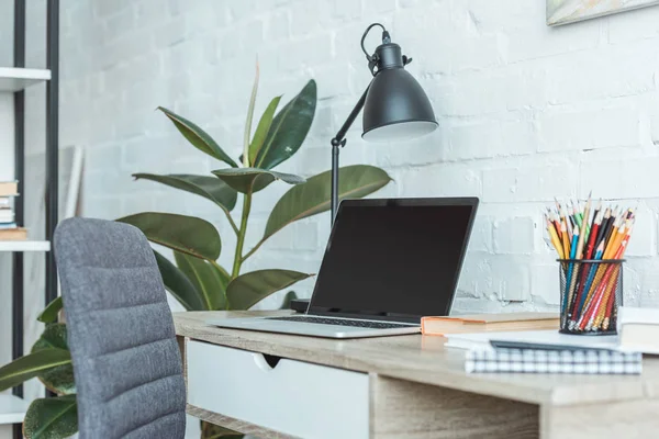 Laptop com tela em branco, livros, candeeiros e lápis na mesa no escritório em casa — Fotografia de Stock