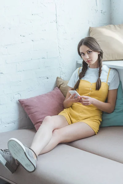 Hermosa chica adolescente con trenzas usando teléfono inteligente y sentado en el sofá - foto de stock