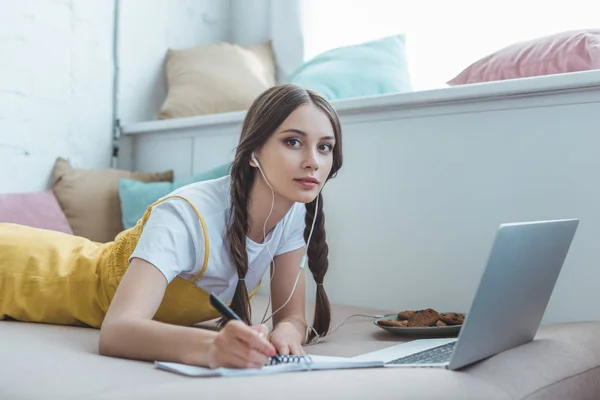 Girl with earphones and laptop writing in copybook — Stock Photo