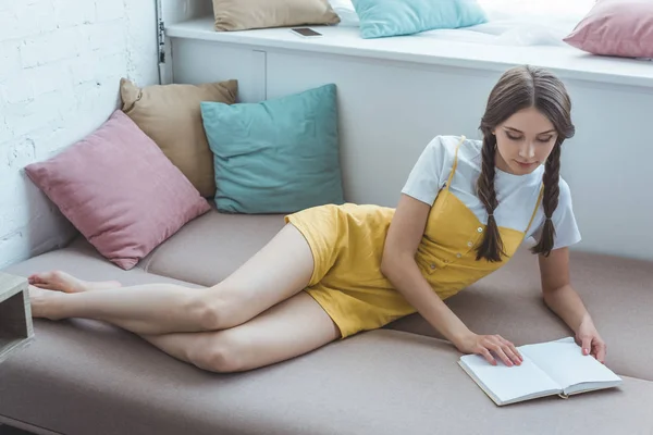 Bela menina adolescente com tranças leitura livro no sofá — Fotografia de Stock