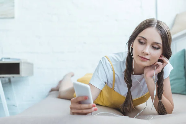 Adolescent fille écoute de la musique avec écouteurs et smartphone tout en étant couché sur canapé — Photo de stock