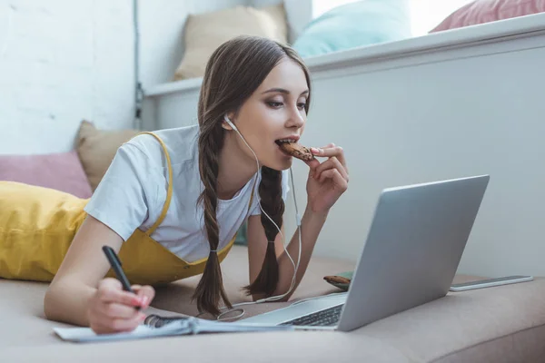 Красивая девочка-подросток ест кулинарные записи в тетрадке и использует ноутбук на диване — стоковое фото