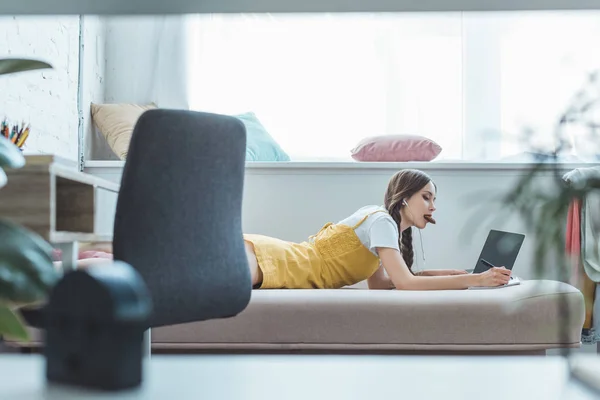 Teen girl eating cookie writing in copybook and using laptop on sofa near window — Stock Photo