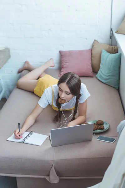 Youth student with cookies using laptop and writing in copybook while lying on sofa at home — Stock Photo