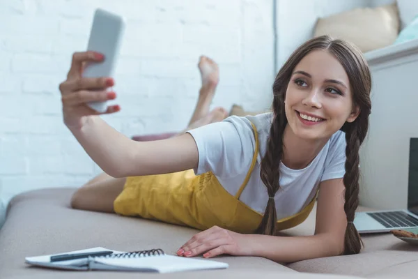 Усміхнена дівчина бере селфі на смартфон, лежачи на дивані з копіювальною книгою та ноутбуком — стокове фото