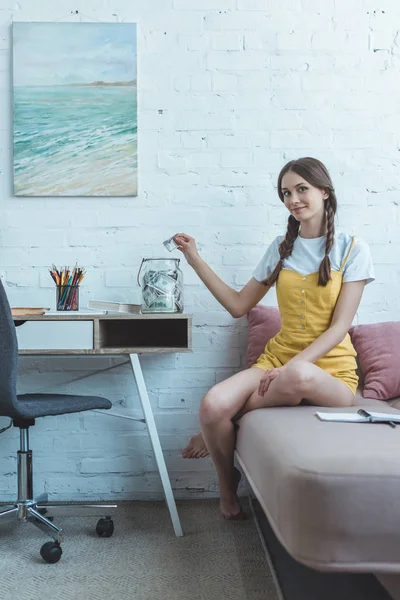 Teenie-Mädchen mit Zöpfen legt Dollarschein in Glasgefäß auf Tisch — Stockfoto