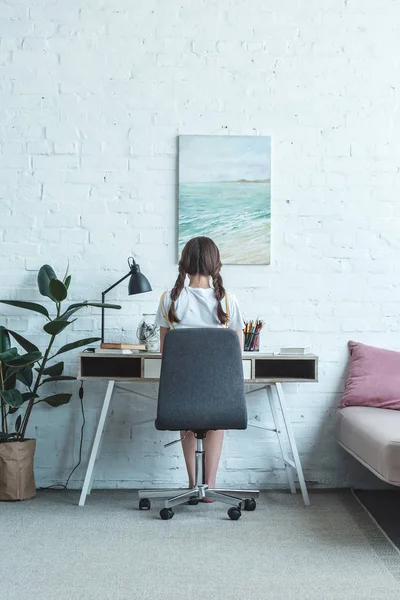Вид ззаду дівчина сидить за столом в кімнаті з картиною на стіні — стокове фото