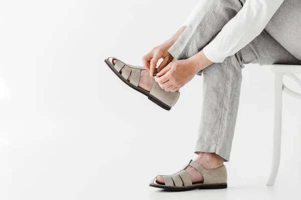 Imagen recortada de modelo masculino en pantalones de lino sentados en silla y poniéndose sandalias aisladas sobre fondo gris - foto de stock