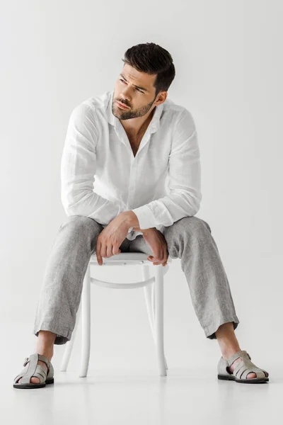 Uomo sicuro di sé in abiti di lino seduto sulla sedia e guardando lontano isolato su sfondo grigio — Foto stock