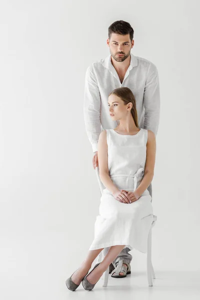 Donna in lino vestito bianco guardando lontano seduto sulla sedia mentre il suo ragazzo in piedi dietro isolato su sfondo grigio — Foto stock