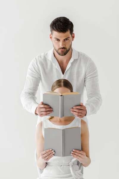 Hombre guapo cubriendo la cara de la novia sentada en la silla y leyendo libro aislado sobre fondo gris - foto de stock