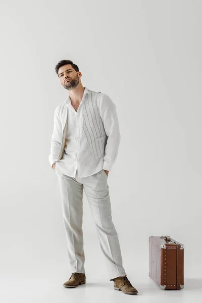 Hombre guapo en ropa de lino posando con las manos en bolsillos cerca de la maleta vintage aislado sobre fondo gris - foto de stock