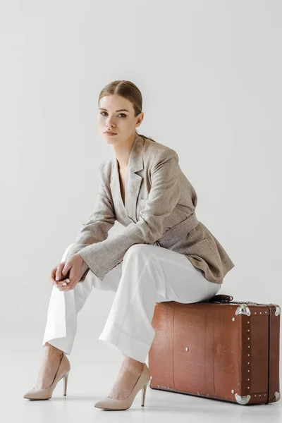 Stylish female traveler in linen jacket sitting on vintage suitcase isolated on grey background — Stock Photo