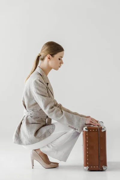 Боковой вид на стильную молодую путешественницу в льняной куртке, открывающую винтажный чемодан, изолированный на сером фоне — стоковое фото