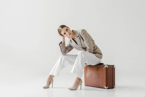 Young stylish female model in linen jacket sitting on vintage suitcase and looking at camera isolated on grey background — Stock Photo