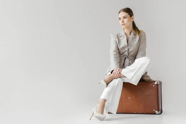 Стильная женщина в льняной куртке сидит на винтажном чемодане и смотрит в сторону изолированы на сером фоне — стоковое фото
