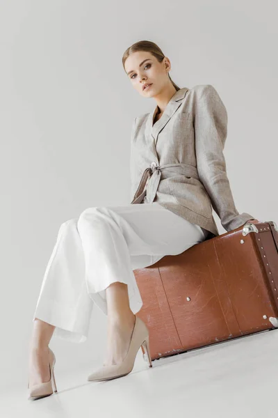 Low angle view of stylish woman in linen jacket sitting on vintage suitcase isolated on grey background — Stock Photo