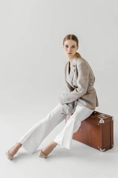 Mulher elegante atraente em jaqueta de linho sentado em mala vintage isolada em fundo cinza — Fotografia de Stock