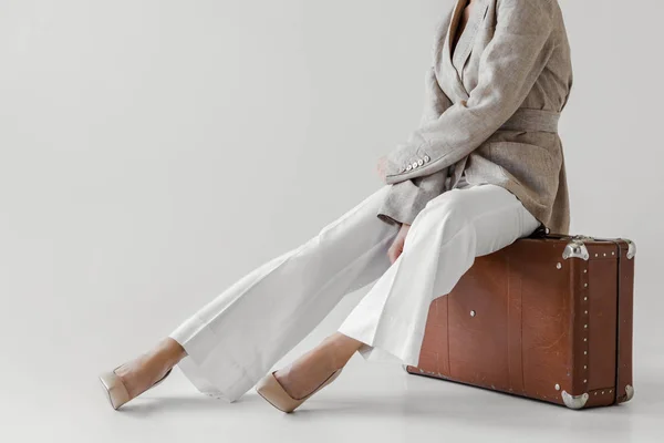Обрезанный образ стильной женщины в льняной куртке, сидящей на винтажном чемодане на сером фоне — стоковое фото
