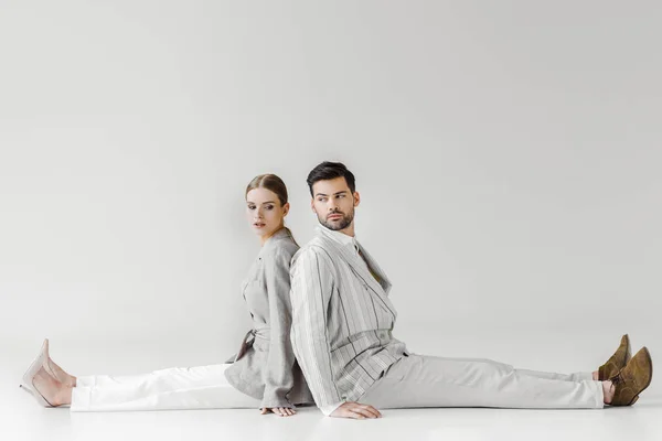 Пара модных моделей в винтажной одежде, сидящих на полу и наклоняющихся спиной к спине на белом — стоковое фото