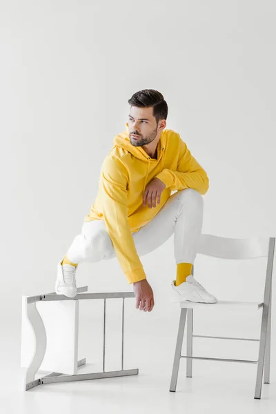 Élégant jeune homme en sweat à capuche jaune assis squats sur des chaises retournées sur blanc — Photo de stock