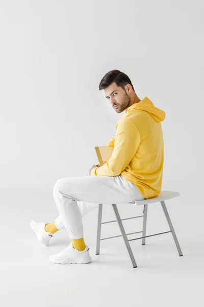 Atractivo joven en sudadera con capucha amarilla sentado en silla volteada en blanco y mirando a la cámara - foto de stock