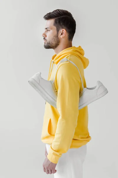 Vue latérale du beau jeune homme en sweat à capuche jaune avec des baskets blanches suspendues à l'épaule isolées sur blanc — Photo de stock