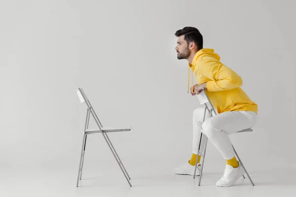 Stylischer junger Mann in gelbem Kapuzenpulli sitzt auf Stuhl vor einem anderen Stuhl auf weißem Grund — Stockfoto