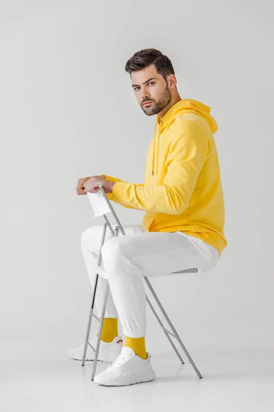 Seitenansicht eines hübschen jungen Mannes in gelbem Kapuzenpulli, der auf einem Stuhl auf weiß sitzt — Stockfoto