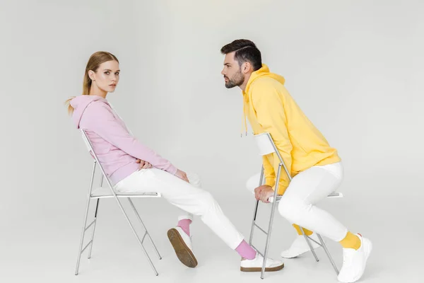 Seitenansicht junger männlicher und weiblicher Models in rosa und gelben Kapuzenpullis, die auf Stühlen auf weißen Stühlen sitzen — Stockfoto