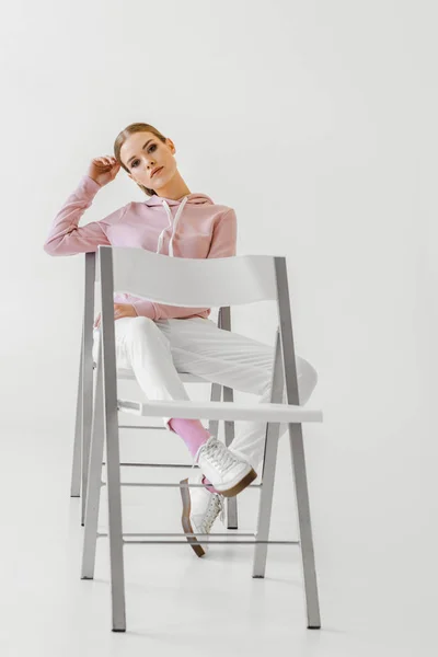 Ernsthafte modische junge Frau sitzt auf Stuhl und blickt in die Kamera auf weiß — Stockfoto