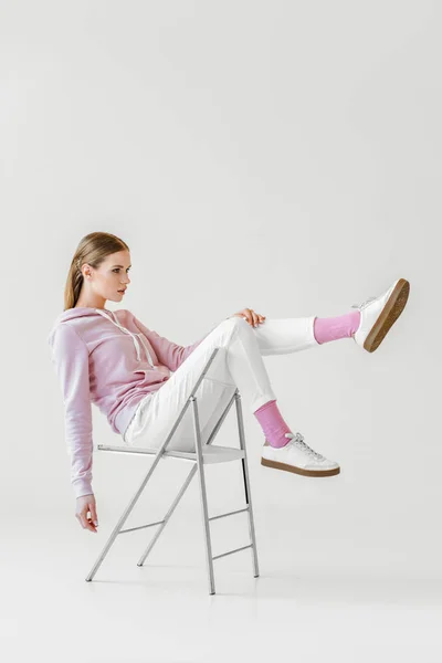 Jeune femme à la mode en sweat à capuche rose assis sur une chaise sur blanc et détournant les yeux — Photo de stock