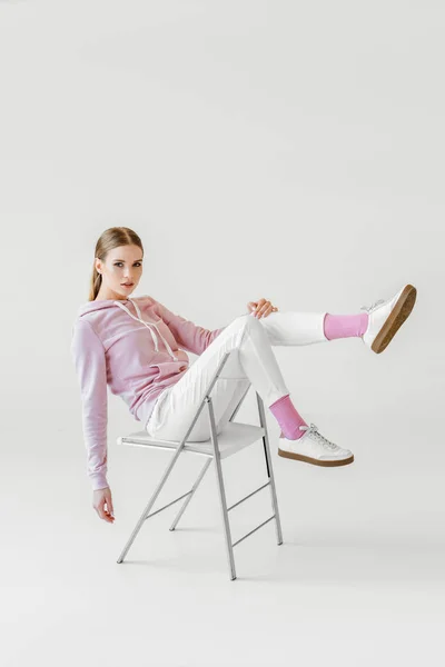 Jovem mulher na moda com capuz rosa sentado na cadeira no branco e olhando para a câmera — Fotografia de Stock