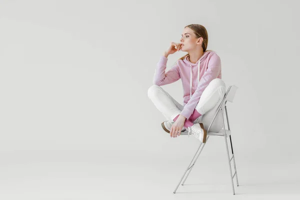 Jeune femme réfléchie en sweat à capuche rose assis sur une chaise sur blanc et détournant les yeux — Photo de stock